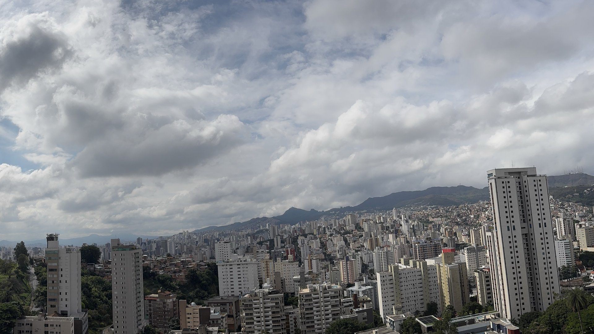 Vista panorâmica de Belo Horizonte a partir do alto do edifício na Av. Raja Gabaglia. Skyline urbano completo do Centro-Sul e do Luxemburgo em primeiro plano, e a silhueta da Serra do Curral atravessando o horizonte sob um céu nublado de meio-dia.