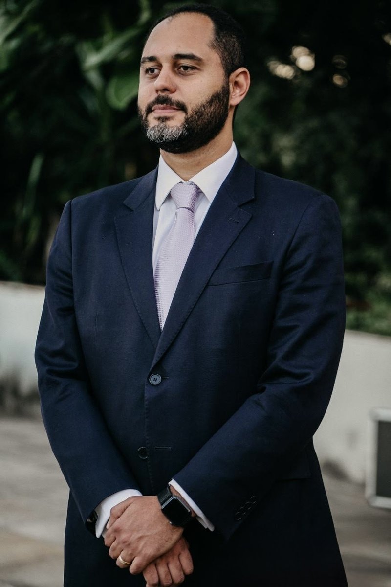 Thiago Alves dos Reis, titular da TR Advocacia, retrato editorial em terno azul-marinho com gravata violeta, ambiente de jardim em segundo plano com folhagem de bananeira.